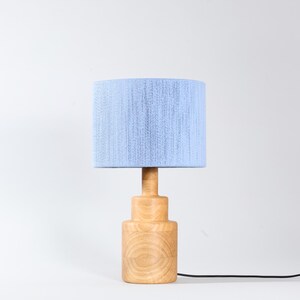 Puede incluir: Una l&aacute;mpara de mesa de madera con una pantalla azul claro. La l&aacute;mpara tiene una base cil&iacute;ndrica y un tallo de madera a juego. La pantalla es de tela azul claro. Un cable de alimentaci&oacute;n negro sale de la base.