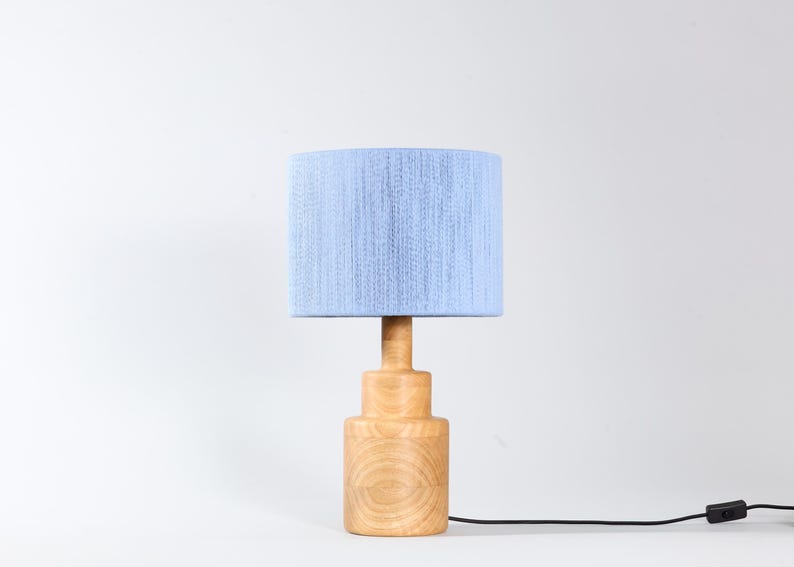 Puede incluir: Una l&aacute;mpara de mesa de madera con una pantalla azul claro. La l&aacute;mpara tiene una base cil&iacute;ndrica y un tallo de madera a juego. La pantalla es de tela azul claro. Un cable de alimentaci&oacute;n negro sale de la base.