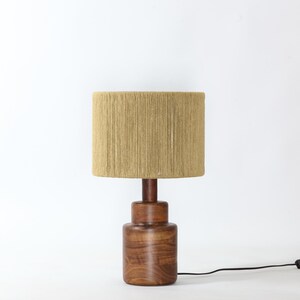 Puede incluir: Una l&aacute;mpara de mesa de madera con una pantalla cil&iacute;ndrica de tela beige. La base es de madera oscura y tiene forma bulbosa. Un cable de alimentaci&oacute;n negro sale de la base.