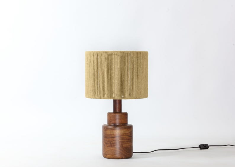Puede incluir: Una l&aacute;mpara de mesa de madera con una pantalla cil&iacute;ndrica de tela beige. La base es de madera oscura y tiene forma bulbosa. Un cable de alimentaci&oacute;n negro sale de la base.