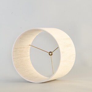 Puede incluir: Una pantalla de l&aacute;mpara cil&iacute;ndrica de color crema con una tela texturizada. La pantalla tiene un dise&ntilde;o circular con un marco de metal color lat&oacute;n. El dise&ntilde;o es simple y moderno, adecuado para varios estilos de interior.