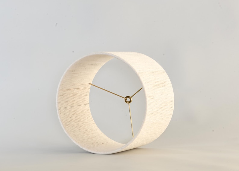 Puede incluir: Una pantalla de l&aacute;mpara cil&iacute;ndrica de color crema con una tela texturizada. La pantalla tiene un dise&ntilde;o circular con un marco de metal color lat&oacute;n. El dise&ntilde;o es simple y moderno, adecuado para varios estilos de interior.