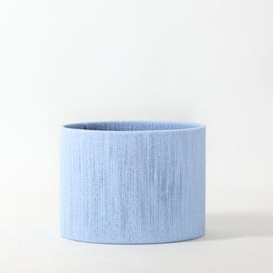 May include: A cylindrical lampshade in a soft, light blue hue. The lampshade has a textured surface, with vertical lines creating a subtle pattern. The lampshade is a simple, yet elegant design, suitable for various home decor styles.