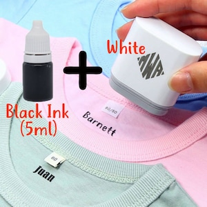May include: A white fabric label maker with a black ink cartridge. The label maker is being used to label a pink shirt with the name "Barnett" and a blue shirt with the name "Juan".