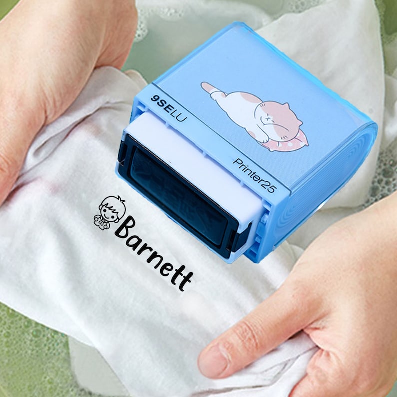 May include: A blue and white self-inking stamp with a cartoon cat design. The stamp reads "9SELU Printer25". The stamp is being used to stamp the name "Barnett" on a white t-shirt.