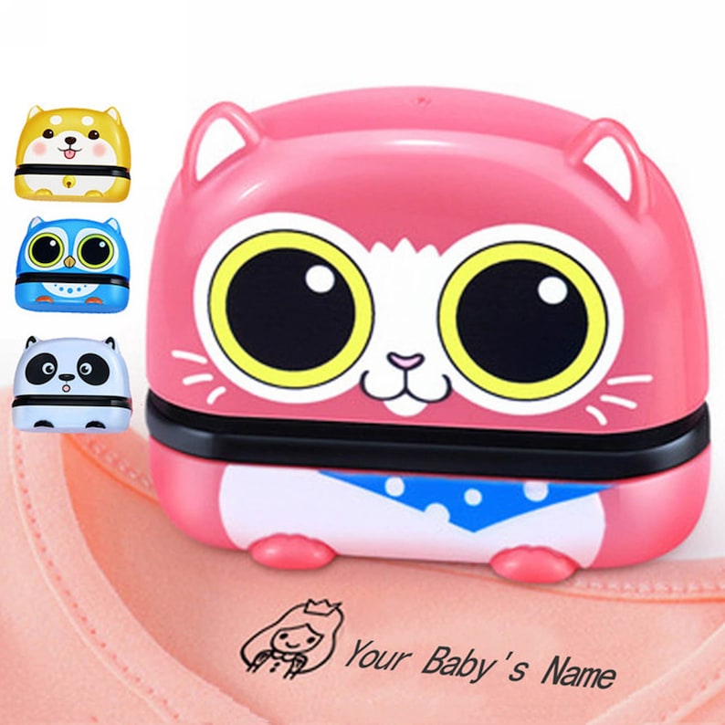 May include: A pink cat-shaped self-inking stamp with large eyes and a blue bandana. The stamp is being used to stamp a pink fabric with the text "Your Baby's Name" and a small illustration of a girl with a crown.