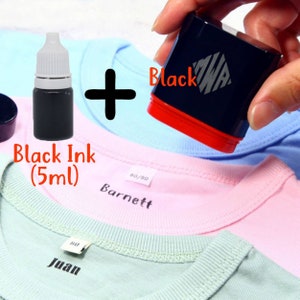 May include: A black ink bottle with a white cap and a black self-inking stamp with the word "Black" printed on it. The stamp is being used to stamp the word "Barnett" on a pink t-shirt. The ink bottle is labeled "Black Ink (5ml)".