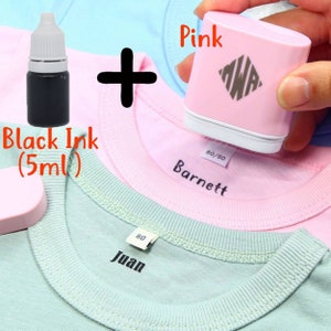 May include: A pink fabric stamper with a black ink bottle. The stamper is being used to stamp the name "Barnett" on a pink shirt. The ink bottle is labeled "Black Ink (5ml)".