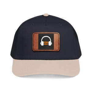 May include: A navy blue baseball cap with a beige brim and a top button. Centered on the front is a rectangular brown leather-like patch featuring a black square with a white headphone icon and the orange text TNTUNES MUSIC printed below it.