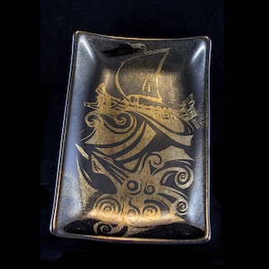 May include: A black rectangular ceramic dish with a gold design of a ship sailing through waves. The design is intricate and detailed, with swirls and curves.