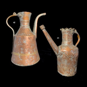 Puede incluir: Dos jarras de cobre antiguas con superficies desgastadas y detalles decorativos. La jarra de la izquierda tiene un pico largo y un asa, mientras que la jarra de la derecha tiene un pico más corto y un asa. La jarra de la izquierda tiene una banda dorada alrededor del medio. Las jarras están etiquetadas como #1 y #2.