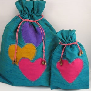 Peut inclure: Deux sacs à cordon de serrage turquoise avec des appliqués en forme de cœur. Le grand sac présente un motif de cœur violet, jaune et rose, tandis que le petit sac a un seul cœur rose. Les deux sacs ont des cordons roses et de petits pompons dorés.