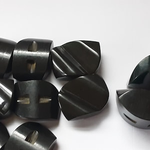 May include: A collection of dark gray buttons with a glossy finish. Some buttons have a curved, ridged design, while others have a more rounded shape with two small rectangular holes. The buttons are arranged on a white surface.