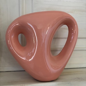 Puede incluir: Un jarrón de cerámica brillante de color coral con un diseño abstracto único. El jarrón presenta dos grandes aberturas circulares y una forma suave y curvada. Es un elemento de decoración del hogar moderno.