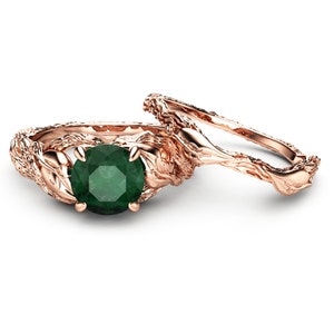 Puede incluir: Un anillo de compromiso de oro rosa con una piedra preciosa de esmeralda verde. La banda del anillo est&aacute; dise&ntilde;ada de forma intrincada con un patr&oacute;n de rama retorcida.