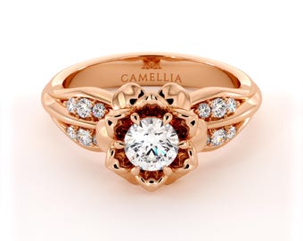 May include: A rose gold engagement ring with a large round diamond set in a flower-shaped setting. The band is adorned with smaller diamonds and the inscription "CAMELLIA" is visible on the band.