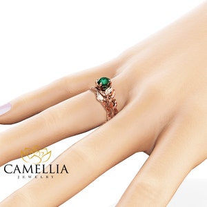 Puede incluir: Anillo de compromiso de oro rosa con una esmeralda verde y un dise&ntilde;o de hojas. El anillo est&aacute; en el dedo de una modelo.