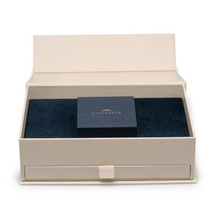Puede incluir: Una caja blanca con un interior de terciopelo azul y una caja negra m&aacute;s peque&ntilde;a con la palabra "CAMELLIA" en letras doradas. La caja tiene un caj&oacute;n.