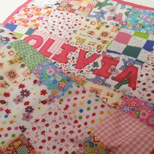 May include: A patchwork quilt featuring a variety of colorful fabrics with floral, polka dot, and teddy bear patterns. The name "OLIVIA" is prominently displayed in large, red, polka-dotted letters. The quilt is trimmed with a red polka dot border.