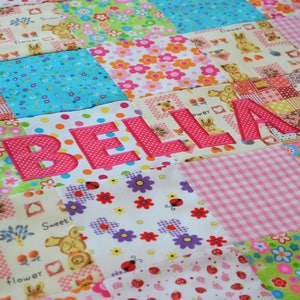May include: A patchwork quilt with a variety of colorful floral, polka dot, and patterned fabrics. The name "BELLA" is appliqu&eacute;d in pink, adding a personalized touch. The quilt is a mix of pink, blue, green, and yellow squares.