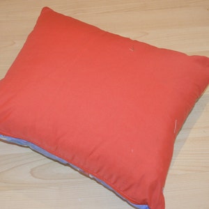 May include: A square pillow with a solid coral-red cover. The pillow's edges reveal a light blue fabric with a floral pattern. The pillow is resting on a light-colored wooden surface.