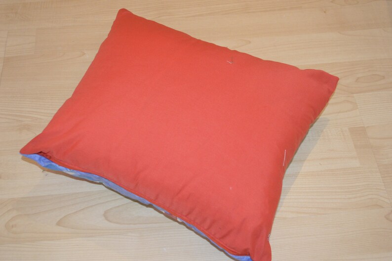 May include: A square pillow with a solid coral-red cover. The pillow's edges reveal a light blue fabric with a floral pattern. The pillow is resting on a light-colored wooden surface.