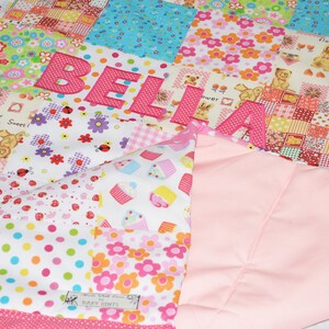 May include: A patchwork quilt with the name "BELIA" appliqued in pink letters. The quilt features various patterned squares with floral, cupcake, and teddy bear designs. A solid pink fabric forms the backing.