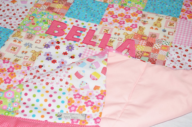 May include: A patchwork quilt with the name "BELIA" appliqued in pink letters. The quilt features various patterned squares with floral, cupcake, and teddy bear designs. A solid pink fabric forms the backing.