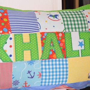 May include: A patchwork pillow with the name "KHALI" appliqued in green polka dot fabric. The pillow features various patterned fabric squares, including red with balloons, blue stripes, and a nautical print. The pillow has a white border and a label that says "Made With Love by Baby Bints."