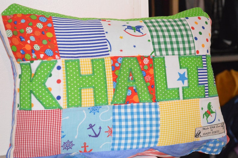 May include: A patchwork pillow with the name "KHALI" appliqued in green polka dot fabric. The pillow features various patterned fabric squares, including red with balloons, blue stripes, and a nautical print. The pillow has a white border and a label that says "Made With Love by Baby Bints."