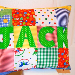 May include: A patchwork pillow with the name "JACK" appliqued in green letters. The pillow features a variety of colorful fabric squares, including polka dots, gingham, and floral patterns. The pillow is rectangular and has a yellow border.