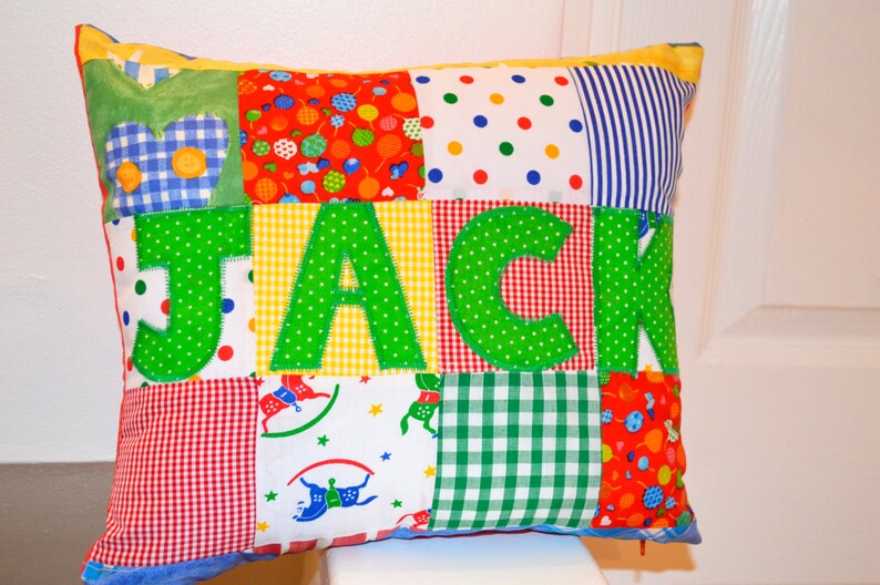 May include: A patchwork pillow with the name "JACK" appliqued in green letters. The pillow features a variety of colorful fabric squares, including polka dots, gingham, and floral patterns. The pillow is rectangular and has a yellow border.