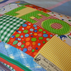 May include: A patchwork pillow with the name "LEO" appliqued in green letters. The pillow features various fabric patterns, including gingham, polka dots, and rocking horse prints. A label reads "Made with love by BABY BIN".