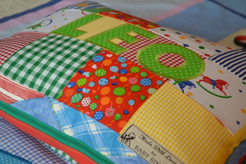 May include: A patchwork pillow with the name "LEO" appliqued in green letters. The pillow features various fabric patterns, including gingham, polka dots, and rocking horse prints. A label reads "Made with love by BABY BIN".