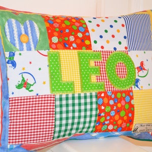 May include: A patchwork pillow with the name "LEO" appliqued in green letters. The pillow features various fabric patterns, including hearts, polka dots, stripes, and gingham. The edges are trimmed with blue fabric and a zipper is visible.