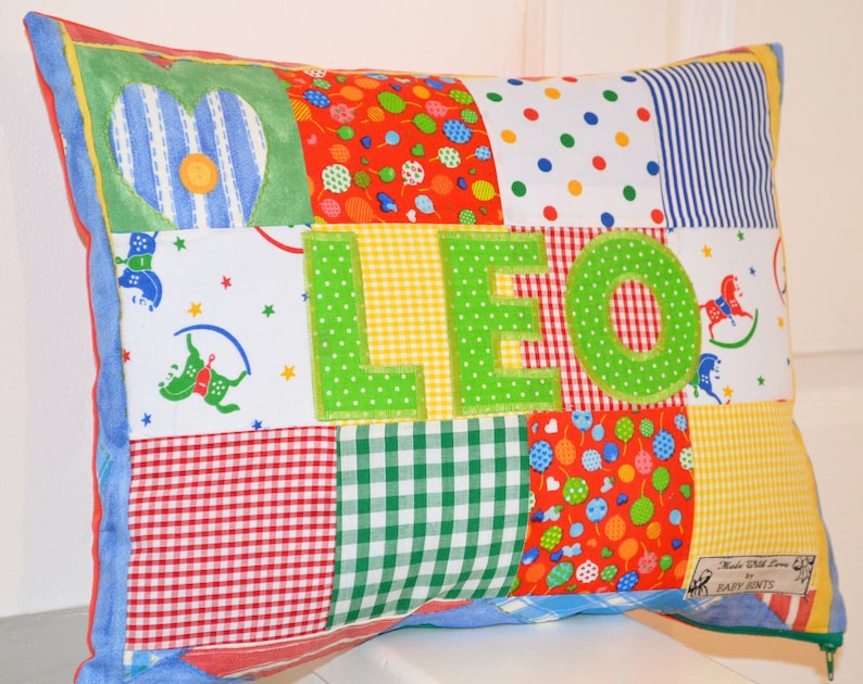May include: A patchwork pillow with the name "LEO" appliqued in green letters. The pillow features various fabric patterns, including hearts, polka dots, stripes, and gingham. The edges are trimmed with blue fabric and a zipper is visible.