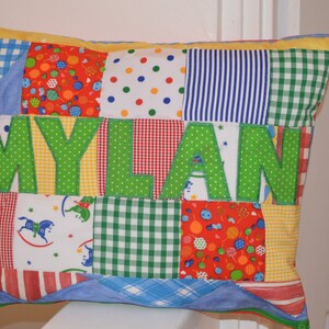 May include: A patchwork pillow with the name "MYLAN" in large green letters. The pillow features various fabric patterns, including gingham, polka dots, and stripes in red, blue, yellow, and green. The pillow is rectangular.