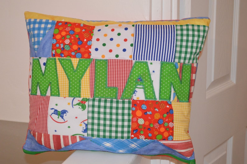 May include: A patchwork pillow with the name "MYLAN" in large green letters. The pillow features various fabric patterns, including gingham, polka dots, and stripes in red, blue, yellow, and green. The pillow is rectangular.