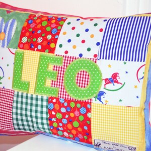 May include: A patchwork pillow with the name "LEO" in green letters. The pillow features various fabric patterns, including polka dots, stripes, and gingham. The edges are trimmed with yellow and blue fabric. The pillow is a colorful and personalized item.