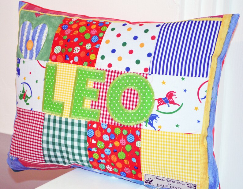May include: A patchwork pillow with the name "LEO" in green letters. The pillow features various fabric patterns, including polka dots, stripes, and gingham. The edges are trimmed with yellow and blue fabric. The pillow is a colorful and personalized item.