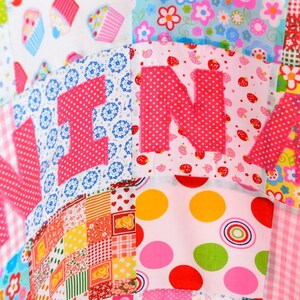 May include: A colorful patchwork quilt with the name "NINA" appliqu&eacute;d in pink polka dot fabric. The quilt features various patterned squares, including floral, polka dot, and gingham designs, in shades of pink, blue, yellow, and green.