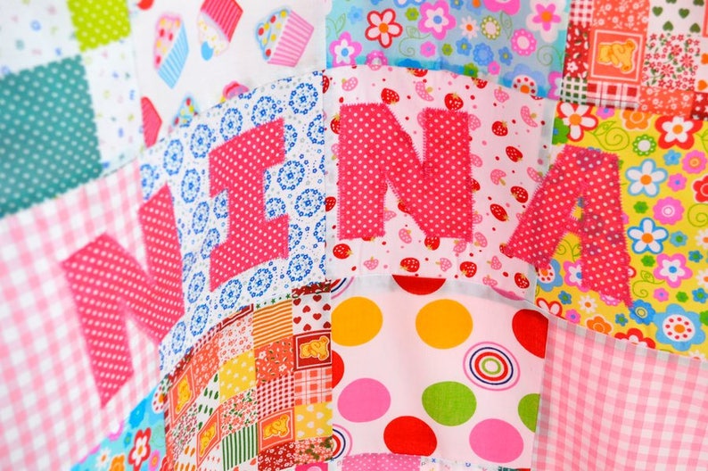 May include: A colorful patchwork quilt with the name "NINA" appliqu&eacute;d in pink polka dot fabric. The quilt features various patterned squares, including floral, polka dot, and gingham designs, in shades of pink, blue, yellow, and green.