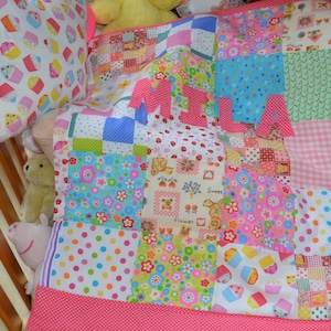 May include: A patchwork baby quilt and pillow set in a crib. The quilt features a variety of colorful patterns, including polka dots, flowers, and cupcakes. The name "MILA" is spelled out in pink letters on the quilt. The pillow has a cupcake pattern.