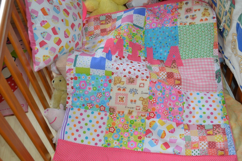 May include: A patchwork baby quilt and pillow set in a crib. The quilt features a variety of colorful patterns, including polka dots, flowers, and cupcakes. The name "MILA" is spelled out in pink letters on the quilt. The pillow has a cupcake pattern.