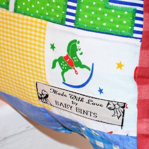 May include: A patchwork textile with a variety of patterns, including gingham, polka dots, and stripes. A label reads "Made With Love by BABY BINTS" and features a green rocking horse illustration. The edges are trimmed with red fabric.
