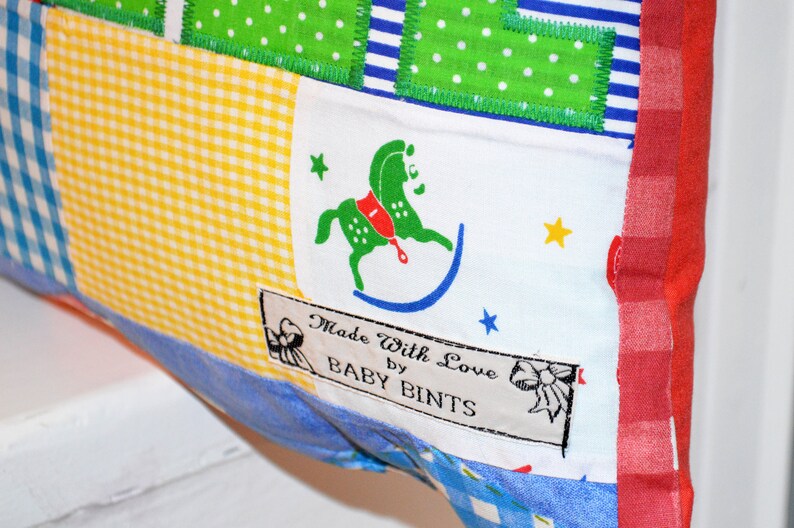 May include: A patchwork textile with a variety of patterns, including gingham, polka dots, and stripes. A label reads "Made With Love by BABY BINTS" and features a green rocking horse illustration. The edges are trimmed with red fabric.