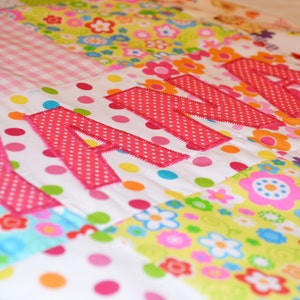 May include: A colorful patchwork quilt with the name "ANNA" appliqu&eacute;d in pink polka dot fabric. The quilt features various floral and polka dot patterns in shades of pink, green, yellow, and blue. The quilt is a handmade item.