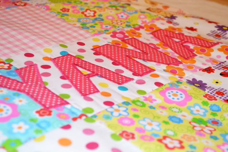 May include: A colorful patchwork quilt with the name "ANNA" appliqu&eacute;d in pink polka dot fabric. The quilt features various floral and polka dot patterns in shades of pink, green, yellow, and blue. The quilt is a handmade item.