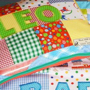 May include: A patchwork pillow with the name "LEO" appliqued in green letters. The pillow features various fabric patterns, including gingham, polka dots, and a train design. The pillow has a green zipper and a label that reads "Made With Love by BABY BINTS."