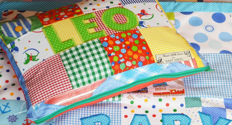 May include: A patchwork pillow with the name "LEO" appliqued in green letters. The pillow features various fabric patterns, including gingham, polka dots, and a train design. The pillow has a green zipper and a label that reads "Made With Love by BABY BINTS."